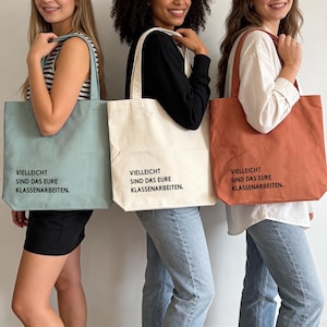 Può includere: Tre borse tote di diversi colori: azzurro, crema e ruggine. Ogni borsa presenta il testo nero: "VIELLEICHT SIND DAS EURE KLASSENARBEITEN." Le borse sono tenute da tre persone. Le borse sono realizzate in tessuto robusto e hanno manici lunghi.