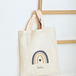 May include: Off-white tote bag featuring a rainbow design in shades of blue, beige, and gold. The bag has the name "Emilia" printed beneath the rainbow. The bag is suspended from a wooden rack, offering a simple, minimalist style.