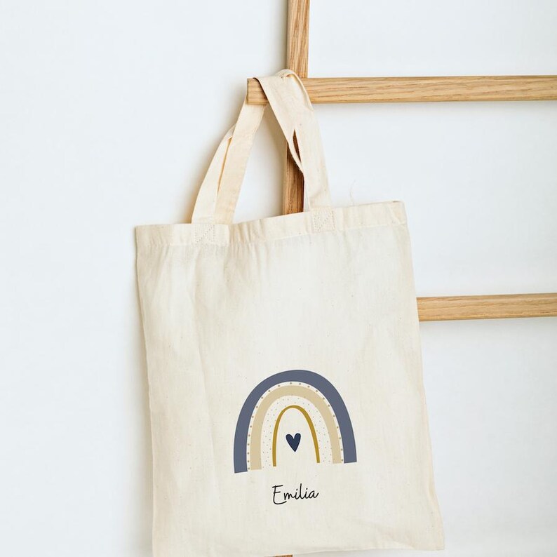 May include: Off-white tote bag featuring a rainbow design in shades of blue, beige, and gold. The bag has the name "Emilia" printed beneath the rainbow. The bag is suspended from a wooden rack, offering a simple, minimalist style.