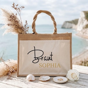 Puede incluir: Un bolso tote rectangular de yute con asa trenzada y bolsillo frontal de lona color crema. El bolsillo presenta el texto en cursiva negra Braut y el texto en bloque dorado SOPHIA. El bolso está decorado con hierba de la pampa y una rosa.