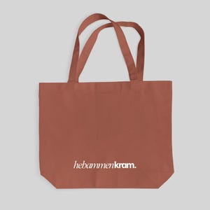 May include: A terracotta-colored tote bag with two shoulder straps. The front of the bag features the white text "hebammenkram." printed in a clean, sans-serif font. The bag has a rectangular shape with a flat bottom and a simple design.