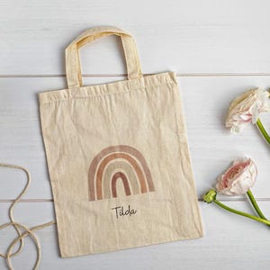 May include: A beige canvas tote bag featuring a brown rainbow design and the name "Tilda". A ball of twine and pink flowers with green stems are arranged on a white wooden surface.