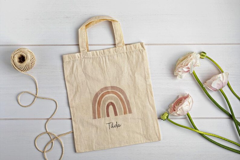 May include: A beige canvas tote bag featuring a brown rainbow design and the name "Tilda". A ball of twine and pink flowers with green stems are arranged on a white wooden surface.