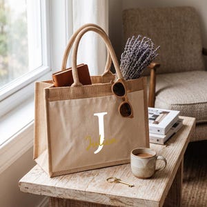 Könnte beinhalten: Eine beigefarbene Jute-Tragetasche mit zwei stabilen Henkeln und einer vorderen Canvas-Tasche. Die Tasche ist mit einem großen weißen Buchstaben J und dem Namen Julien in goldener Schreibschrift bedruckt. Sie enthält ein braunes Leder-Notizbuch.