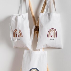 May include: Three white canvas tote bags with rainbow designs and personalised names. The bags are hanging on a wooden rack. The rainbow colours include shades of brown, burgundy, and navy blue. Each bag has a different name: Tilda, Sophia, and Emilia.