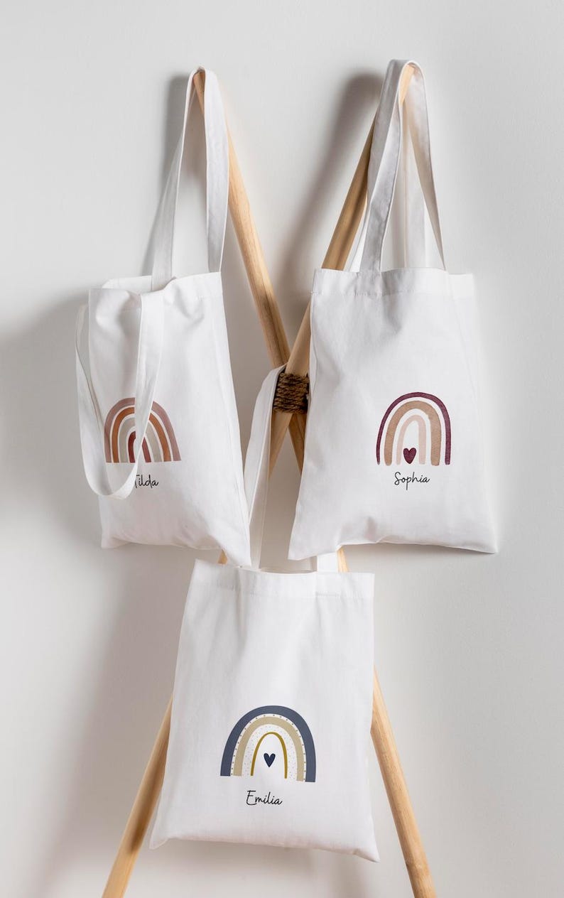 May include: Three white canvas tote bags with rainbow designs and personalised names. The bags are hanging on a wooden rack. The rainbow colours include shades of brown, burgundy, and navy blue. Each bag has a different name: Tilda, Sophia, and Emilia.