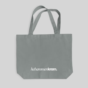 May include: A grey tote bag featuring two shoulder straps and the white text "hebammenkram." printed in a serif font near the bottom center. The bag is constructed from a solid grey fabric with a rectangular shape and a flat base.