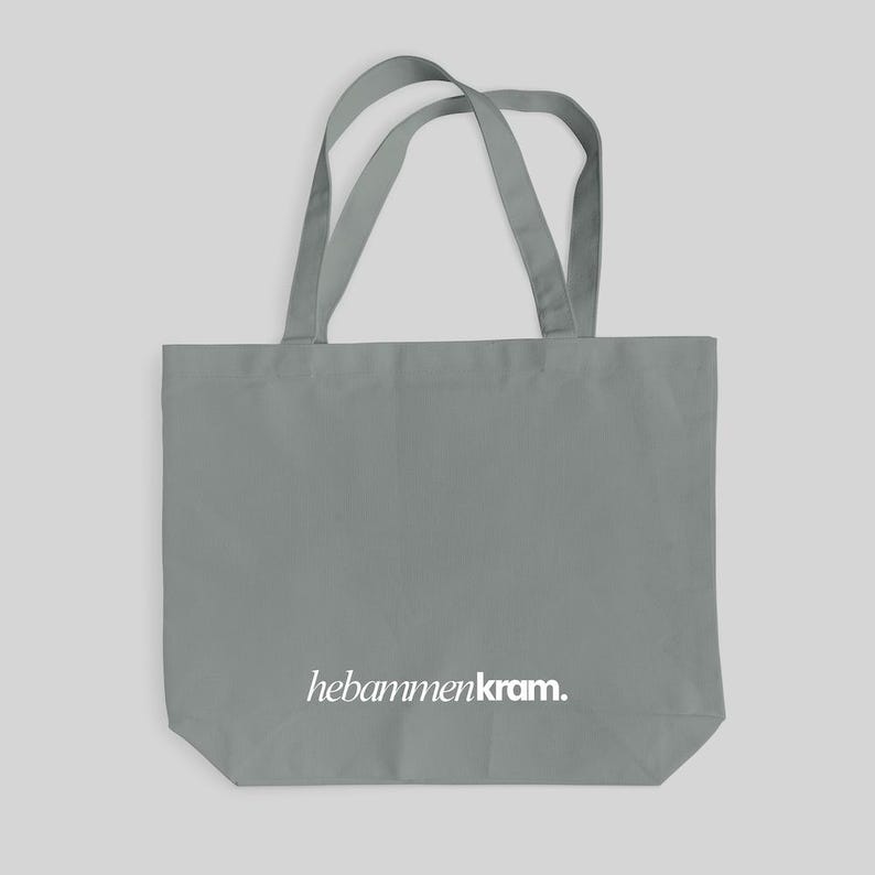 May include: A grey tote bag featuring two shoulder straps and the white text "hebammenkram." printed in a serif font near the bottom center. The bag is constructed from a solid grey fabric with a rectangular shape and a flat base.