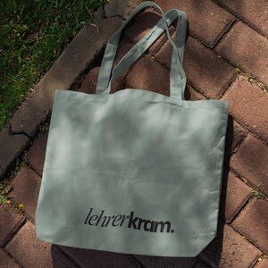 May include: A sage green tote bag with long handles, resting on a brick surface with grass. The bag features the text "lehrerkram." in black, centered on the front. The bag is made of a soft, durable material.