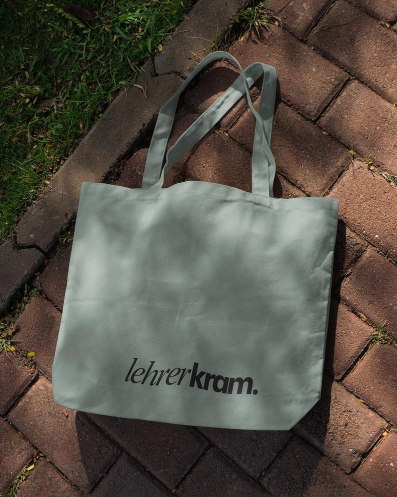 May include: A sage green tote bag with long handles, resting on a brick surface with grass. The bag features the text "lehrerkram." in black, centered on the front. The bag is made of a soft, durable material.