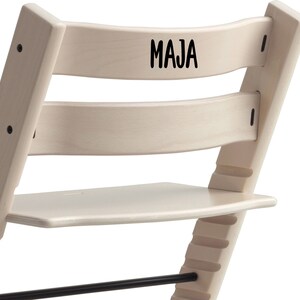 Puede incluir: Una silla de madera blanca con un reposapi&eacute;s negro y el nombre "MAJA" escrito en el respaldo.