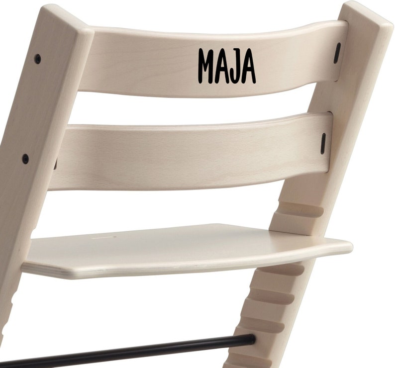 Puede incluir: Una silla de madera blanca con un reposapi&eacute;s negro y el nombre "MAJA" escrito en el respaldo.