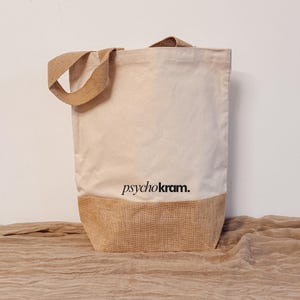 Könnte beinhalten: Eine cremefarbene Canvas-Tragetasche mit zwei beigen Jutehenkeln und einem passenden Juteboden. Auf der Vorderseite der Tasche befindet sich der schwarze Schriftzug "psychokram." in einer kleingeschriebenen Serifenschrift über dem Boden.