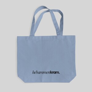 May include: A light blue tote bag with two matching shoulder straps. The bag features the text "hebammenkram." printed in a black, serif-style font centered near the bottom edge of the front panel. The design is simple and functional.