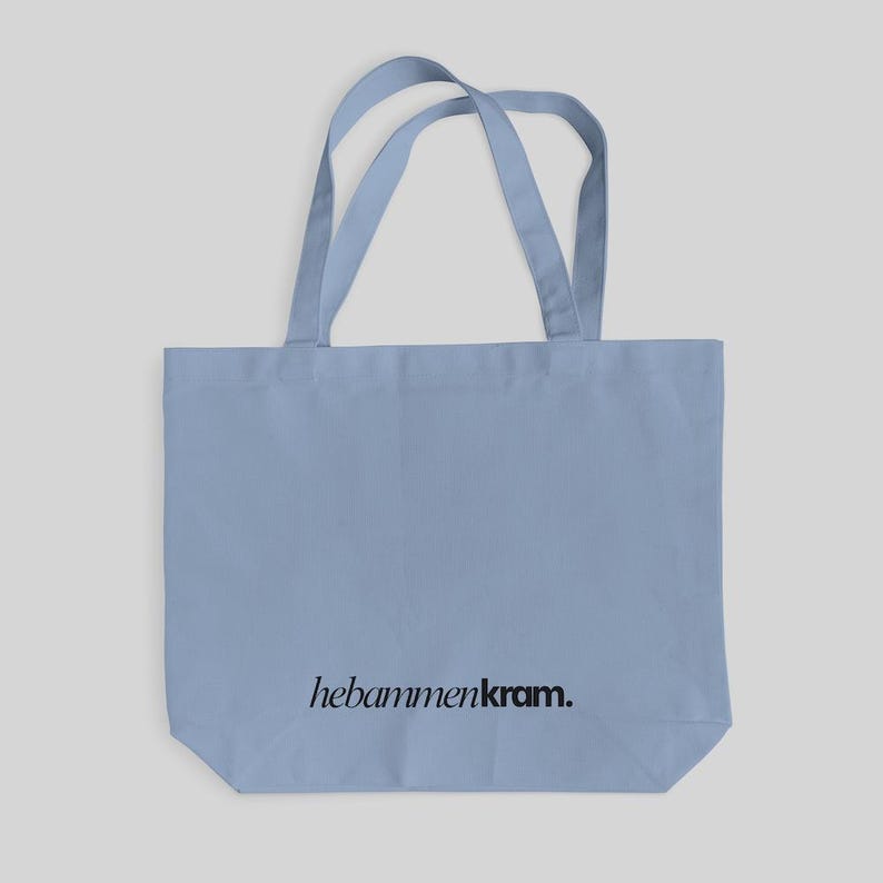 May include: A light blue tote bag with two matching shoulder straps. The bag features the text "hebammenkram." printed in a black, serif-style font centered near the bottom edge of the front panel. The design is simple and functional.