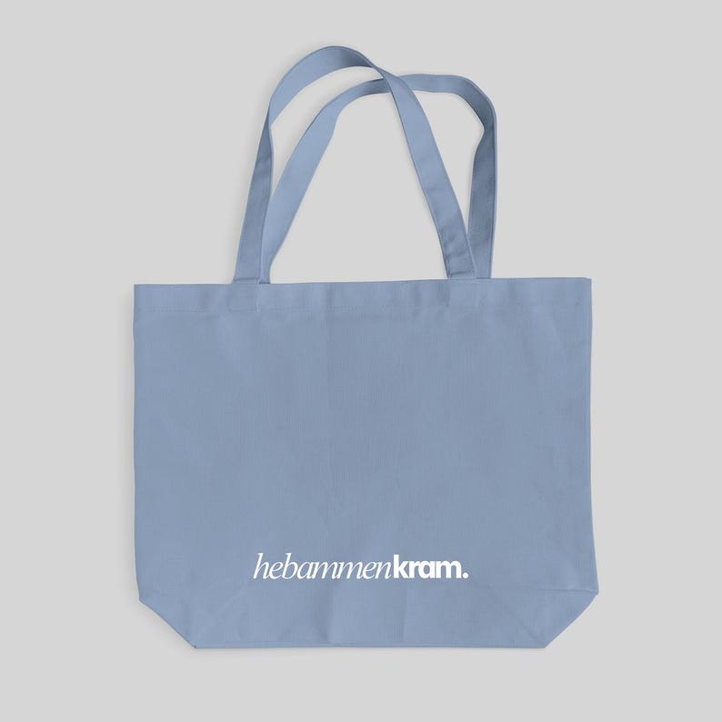 May include: A light blue tote bag with two shoulder straps and the white text hebammenkram. printed in a lowercase serif font on the lower front section. The bag features a rectangular body with a flat base and a simple, functional design.