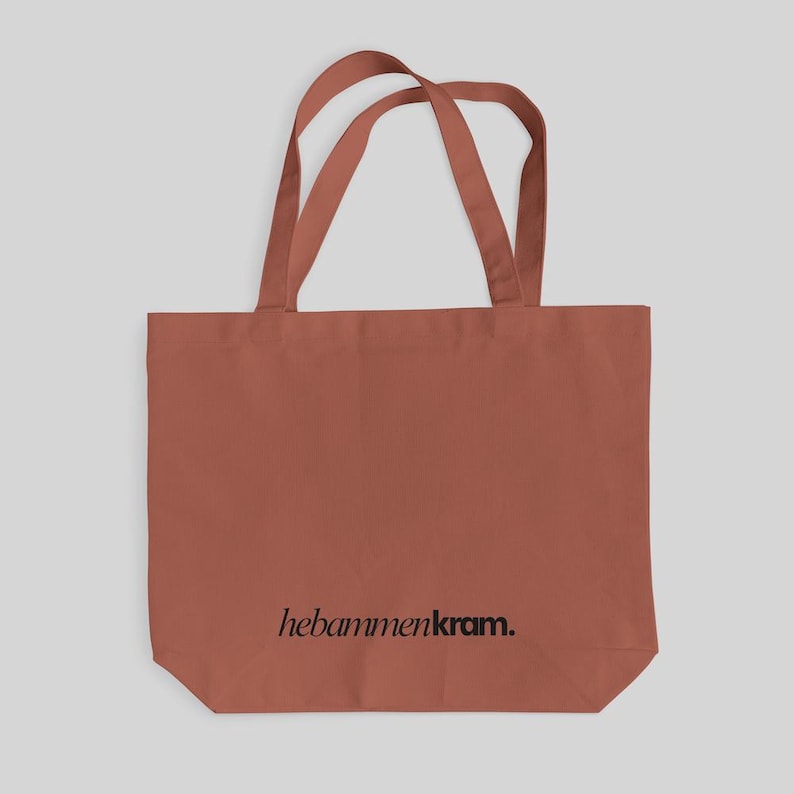 May include: A terracotta-colored tote bag with two shoulder straps. The bag features the text "hebammenkram." printed in a black, lowercase serif font centered near the bottom. The design is simple, with a rectangular body and flat base.