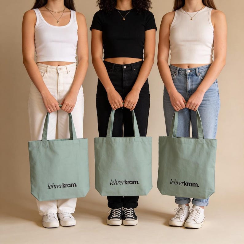 May include: Three people holding sage green tote bags with the text "lehrerkram." printed in black. The bags are rectangular with handles. The people are wearing casual outfits, including jeans, white pants, and black pants.