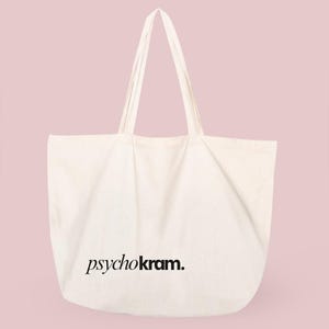 Może przedstawiać: Kremowa torba typu tote z płótna z dwoma długimi paskami na ramię. Z przodu torby znajduje się napis "psychokram." wydrukowany pogrubioną, czarną czcionką szeryfową małymi literami. Torba ma szeroki, prostokątny korpus z płaskim dnem.