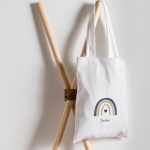 May include: White tote bag featuring a rainbow design in blue, gold, and a small black heart. The name "Emilia" is printed below the rainbow. The bag is suspended on a wooden rack.