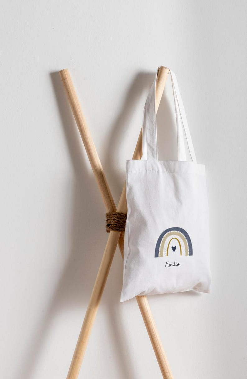 May include: White tote bag featuring a rainbow design in blue, gold, and a small black heart. The name "Emilia" is printed below the rainbow. The bag is suspended on a wooden rack.