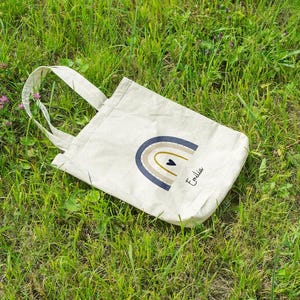 May include: A cream tote bag featuring a rainbow design in blue, tan, and gold, with a small black heart. The name "Emilia" is written in script. The bag rests on green grass.