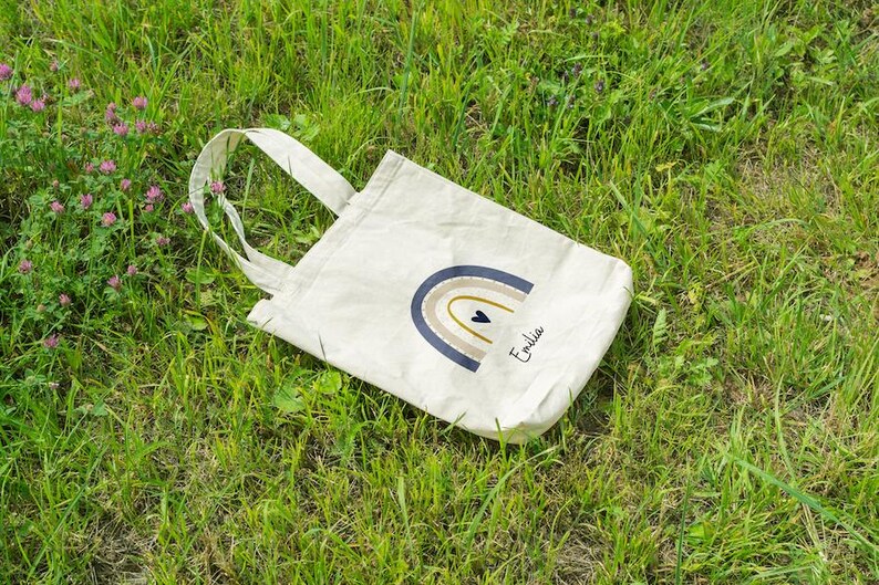 May include: A cream tote bag featuring a rainbow design in blue, tan, and gold, with a small black heart. The name "Emilia" is written in script. The bag rests on green grass.