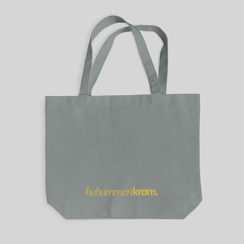 May include: A grey tote bag featuring two shoulder straps and the text hebammenkram. printed in yellow on the lower front section. The bag is constructed from a solid grey fabric with a rectangular shape and a flat base for storage.