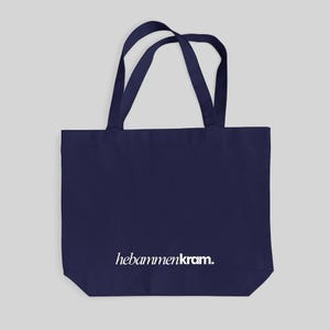 May include: A dark navy blue tote bag featuring two shoulder straps and the white text hebammenkram. printed in a lowercase serif font across the lower center portion of the bag. The bag has a rectangular shape with a flat bottom base.