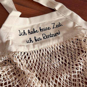 Könnte beinhalten: Eine cremefarbene Baumwoll-Tragetasche mit einem Netz-Körper und zwei langen Henkeln. Der obere Teil aus festem Stoff ist mit dem schwarzen Text bedruckt: Ich habe keine Zeit. ich bin Rentner! Die Tasche hat eine gewebte Struktur und ist naturweiß.