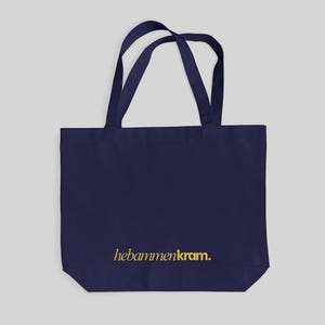 May include: A dark navy blue tote bag with two matching shoulder straps. The front of the bag features the text "hebammenkram." printed in a gold-colored font near the bottom center. The bag has a rectangular shape and a flat base.