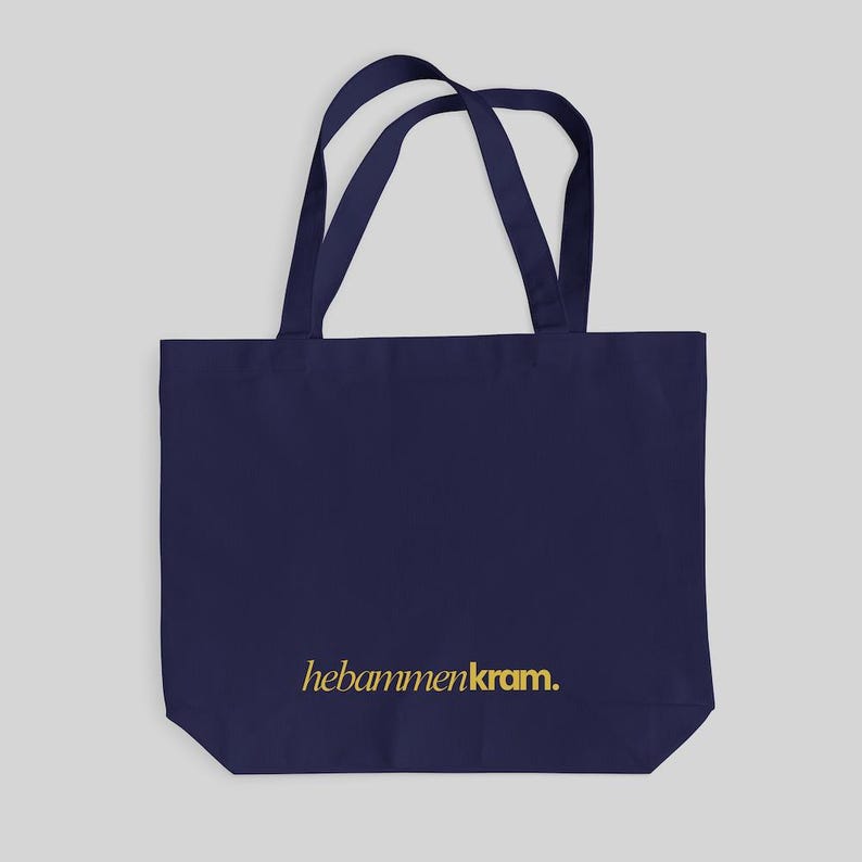 May include: A dark navy blue tote bag with two matching shoulder straps. The front of the bag features the text "hebammenkram." printed in a gold-colored font near the bottom center. The bag has a rectangular shape and a flat base.