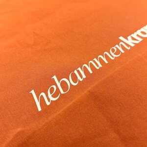 May include: A bright orange fabric tote bag featuring the white text hebammenkro printed diagonally across the surface. The material has a visible woven texture and the lettering is rendered in a clean, sans-serif font style.