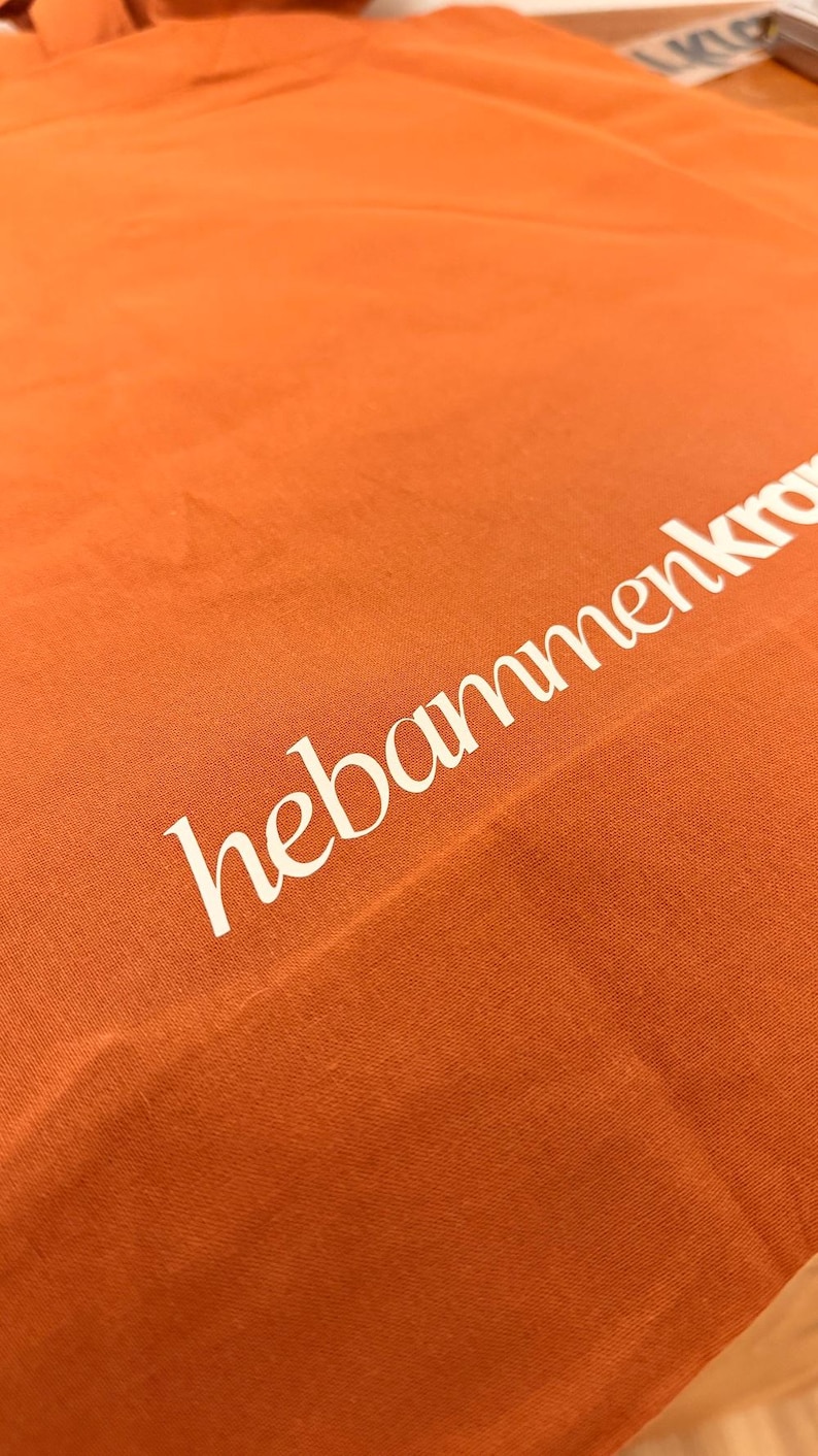 May include: A bright orange fabric tote bag featuring the white text hebammenkro printed diagonally across the surface. The material has a visible woven texture and the lettering is rendered in a clean, sans-serif font style.