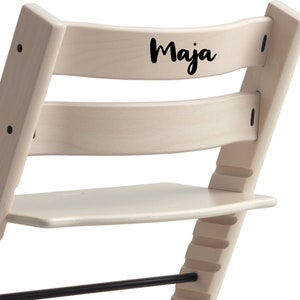 Puede incluir: Una silla de madera blanca con un reposapi&eacute;s negro y una etiqueta de nombre negra en la parte posterior que dice "Maja".