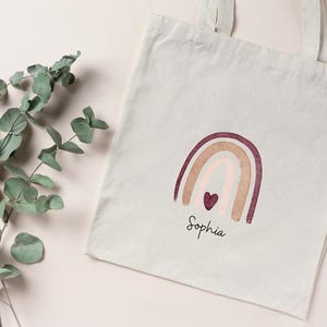 May include: A cream-coloured tote bag featuring a rainbow design in burgundy, beige, and pink, with a small heart. The name "Sophia" is written below the rainbow in a script font. A sprig of eucalyptus is on the left.