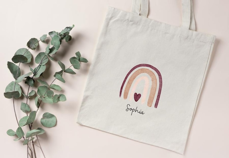 May include: A cream-coloured tote bag featuring a rainbow design in burgundy, beige, and pink, with a small heart. The name "Sophia" is written below the rainbow in a script font. A sprig of eucalyptus is on the left.