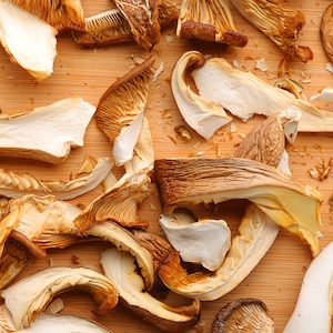 May include: Overhead shot of dried oyster mushrooms on a light brown wooden surface. The mushrooms vary in color from light tan to dark brown, with a variety of shapes and sizes. Some are whole, while others are broken into pieces.