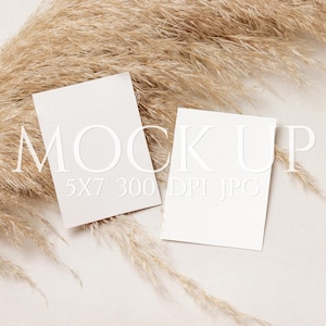 Puede incluir: Dos tarjetas rectangulares blancas con la palabra "MOCKUP" en relieve y el texto "5x7 300 DPI JPG". Las tarjetas están dispuestas sobre una superficie beige con hierba ornamental seca de color marrón claro.