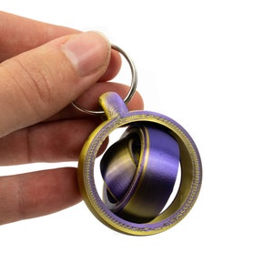 Puede incluir: Un llavero impreso en 3D con un diseño circular. El llavero presenta un anillo exterior de color dorado y un diseño interior morado y dorado. El llavero se sujeta con un llavero plateado.