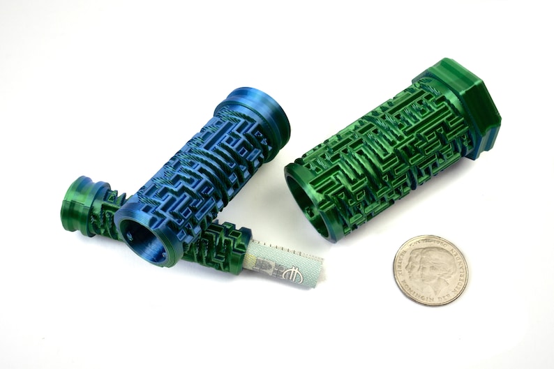 May include: Three 3D-printed cylindrical containers with a maze-like design in shades of blue and green. One container holds a rolled-up bill. A coin is next to the containers. The objects are on a white surface.