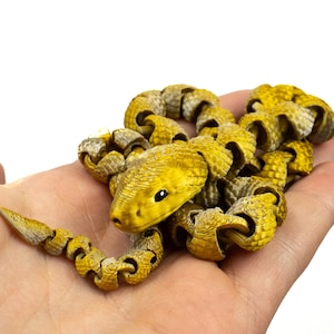 Op de afbeelding: Een gele en bruine slangenfiguur met een gedetailleerd, gestructureerd ontwerp. De slang is opgerold en wordt in een open hand tegen een witte achtergrond gehouden. De kop van de slang is zichtbaar, met een zwart oog.