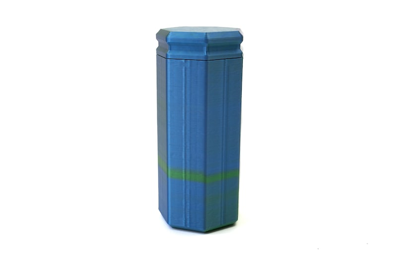 May include: A blue, hexagonal-shaped container with a matching lid. The container has a textured surface and a green stripe around the middle. The design is reminiscent of a pillar or column.