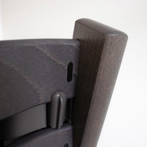 May include: A dark gray wooden high chair with a black plastic strap and a black plastic buckle. The chair has a simple design and is made of high-quality materials.