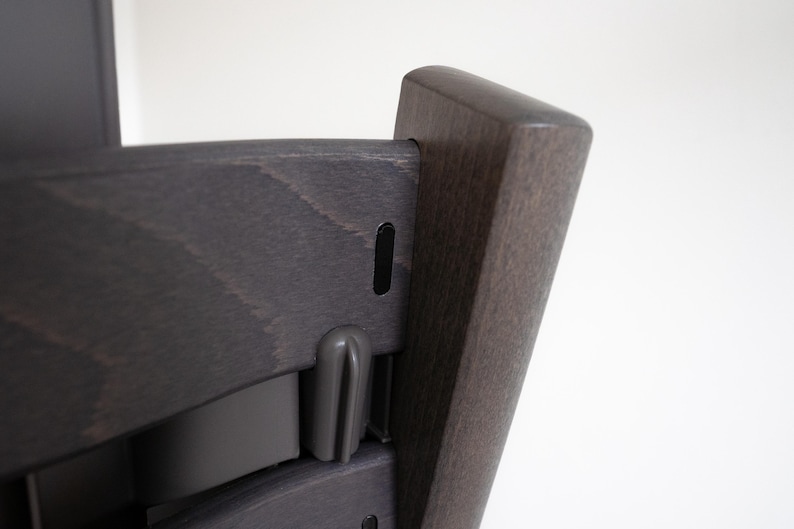 May include: A dark gray wooden high chair with a black plastic strap and a black plastic buckle. The chair has a simple design and is made of high-quality materials.