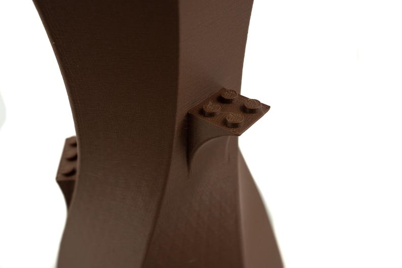 Peut inclure: Un vase en c&eacute;ramique brun au design moderne et unique. Le vase a une surface textur&eacute;e et pr&eacute;sente une petite ouverture carr&eacute;e en haut. Le vase est d&eacute;cor&eacute; d'une petite pi&egrave;ce en plastique de type Lego brune sur le c&ocirc;t&eacute;.