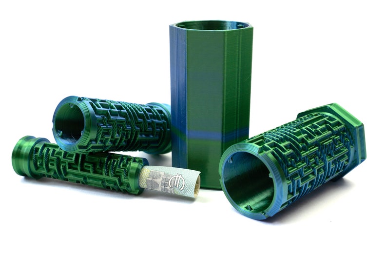 May include: A collection of green and blue 3D-printed puzzle containers. The containers have a maze-like design and cylindrical shapes. One container holds a rolled-up paper item. The objects are on a white background.
