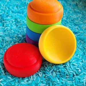 Può includere: Una collezione di giocattoli in legno colorati, tra cui una pila di pezzi rotondi arancioni, verdi, blu e viola, più un giocattolo rosso e uno giallo a forma di ciotola. I giocattoli sono su riso blu.