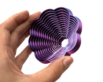 Puede incluir: Una escultura abstracta en capas, en tonos morados y azules, sostenida en una mano. El objeto tiene forma de flor con capas concéntricas y curvas. El color cambia de morado a azul, creando un brillo metálico. El fondo es blanco liso.