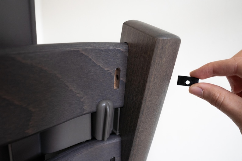 May include: A dark gray wooden chair with a black plastic piece being held by a hand. The chair has a hole in the back of the seat.